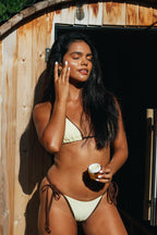 A woman wearing a cream-colored string bikini stands in front of a rustic wooden structure, applying HGO Face & Eye Creme with one hand while holding a Face & Eye Crème jar in the other.